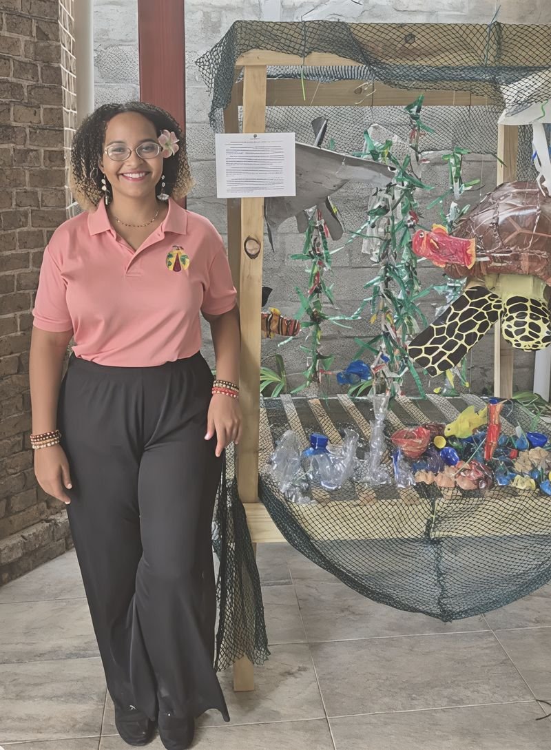 A smiling young woman in a 195in365 polo shirt standing beside an ocean sculpture made from recycled plastic, with a sea turtle, fish and seaweed hanging in a fishing net